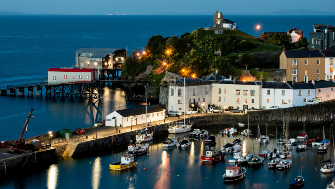 Tenby Harbour at night. Moored boats in the foreground are reflected in the smooth water. The castle ruins are at the top of the hill.