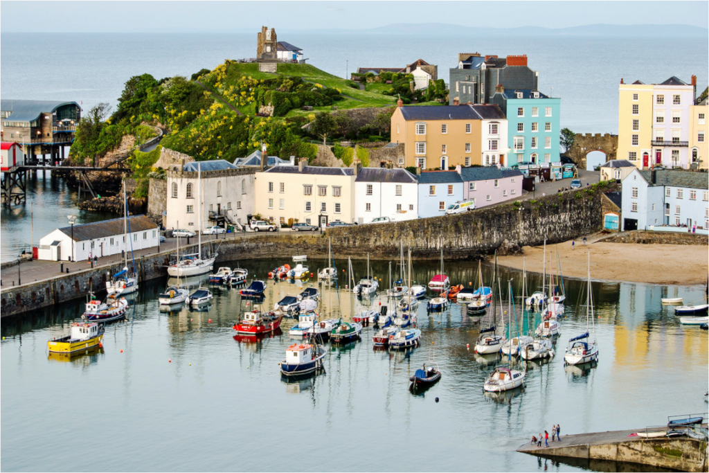 Tenby Harbour. The tide is in and several sailing yachts and fishing boats are swinging on their moorings. The remains of Tenby Castle can be seen on the hilltop behind the harbour.