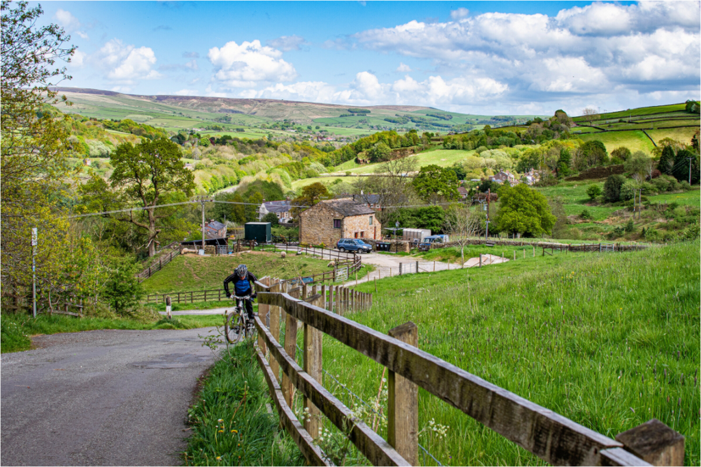 A man cycling Uphill towards along a country lane with a small cottage in the background.