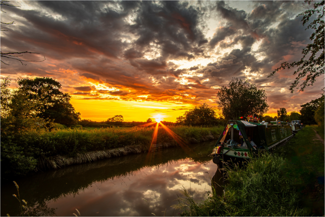 A sunset scene with sun rays extending over a canal in the foreground and a moored narrowboat.