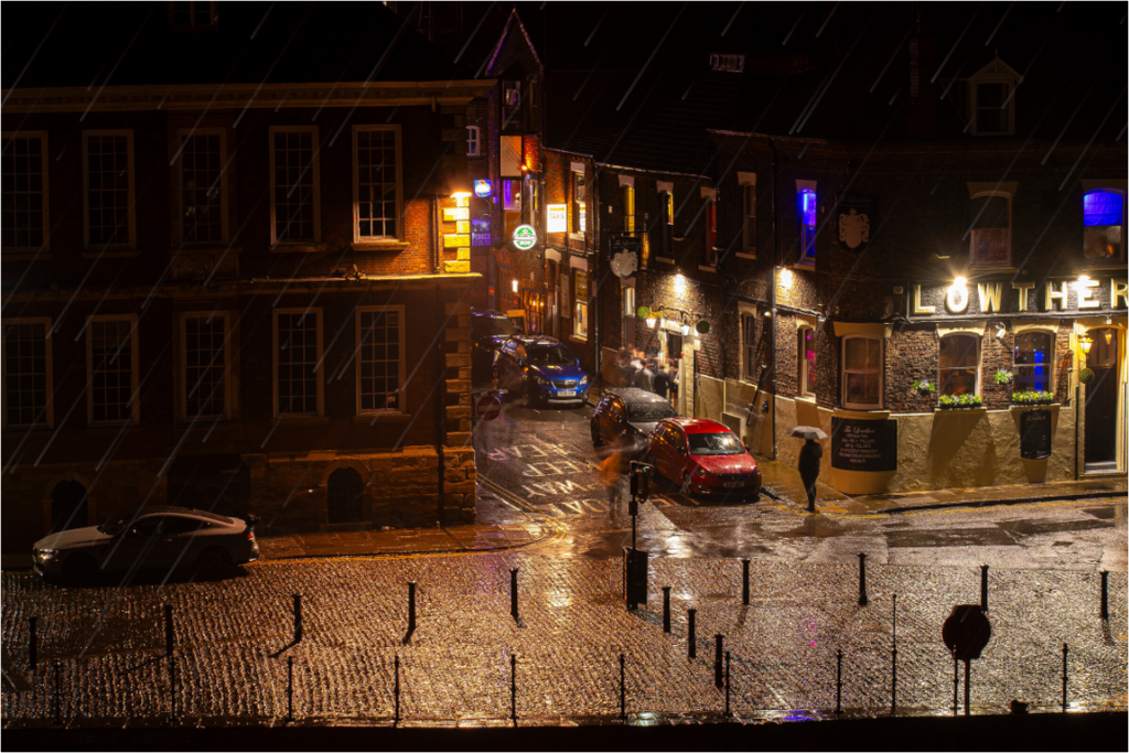 York street on a wet night with illuminated buildings, cars on a cobblestone street and rain.